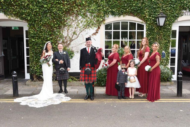 A bride, bagpiper, and wedding party in burgundy dresses posing outside a greenery-covered venue